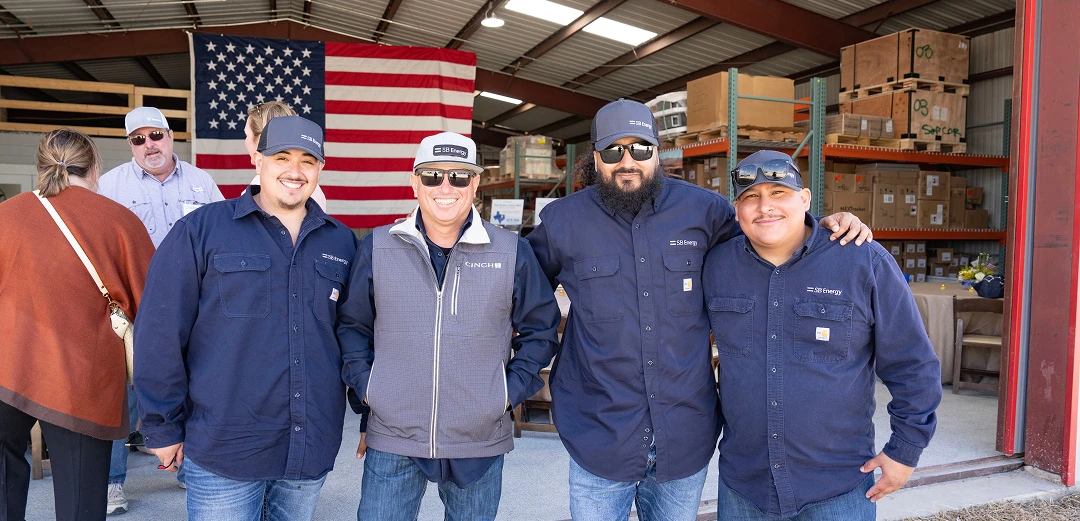 Four SB Energy workers wearing blue shirts standing in front of an American flag inside a warehouse.